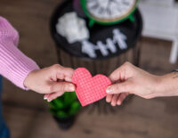 Двама души държат едната страна на текстурирано червено сърце. Малка масичка на заден план държи часовник и хартиени изрезки на хора, подсказващи теми за време и взаимоотношения. Сдружение 'Бъдеще за децата'